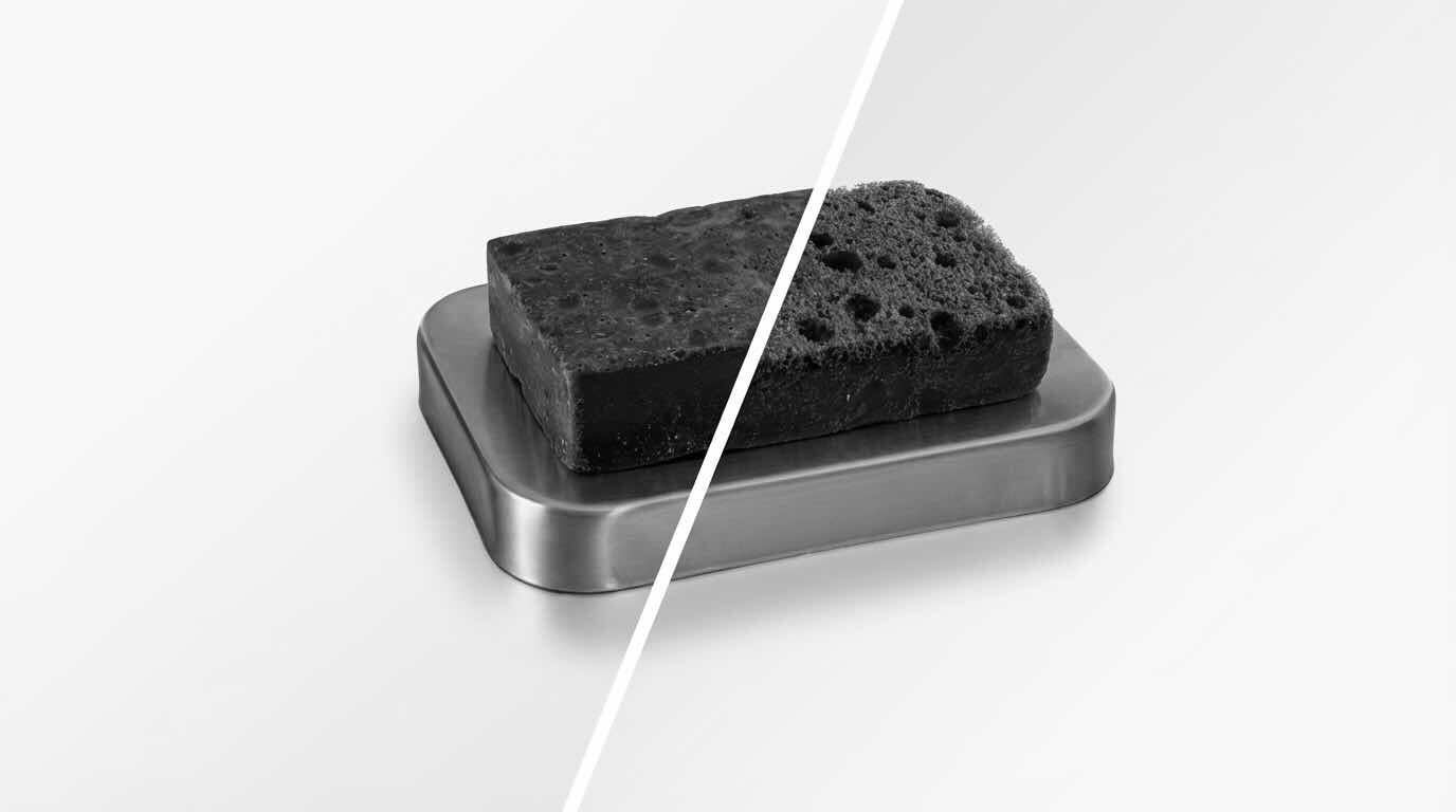 Ein eckiger, poröser Schwamm liegt auf einem silberfarbenen Behälter. Das Bild ist in Graustufen gehalten, mit einer diagonalen Linie, die den Schwamm in zwei Hälften teilt.
