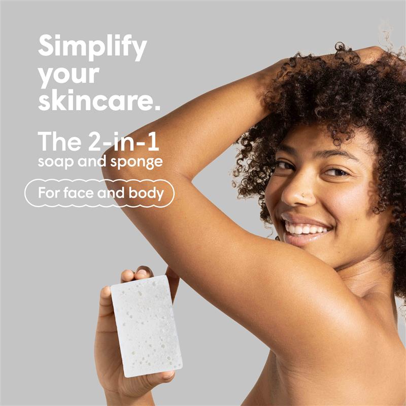Lächelnde Frau mit lockigem Haar hält einen weißen Badeschwamm vor grauem Hintergrund. Text: "Simplify your skincare. The 2-in-1 soap and sponge For face and body".