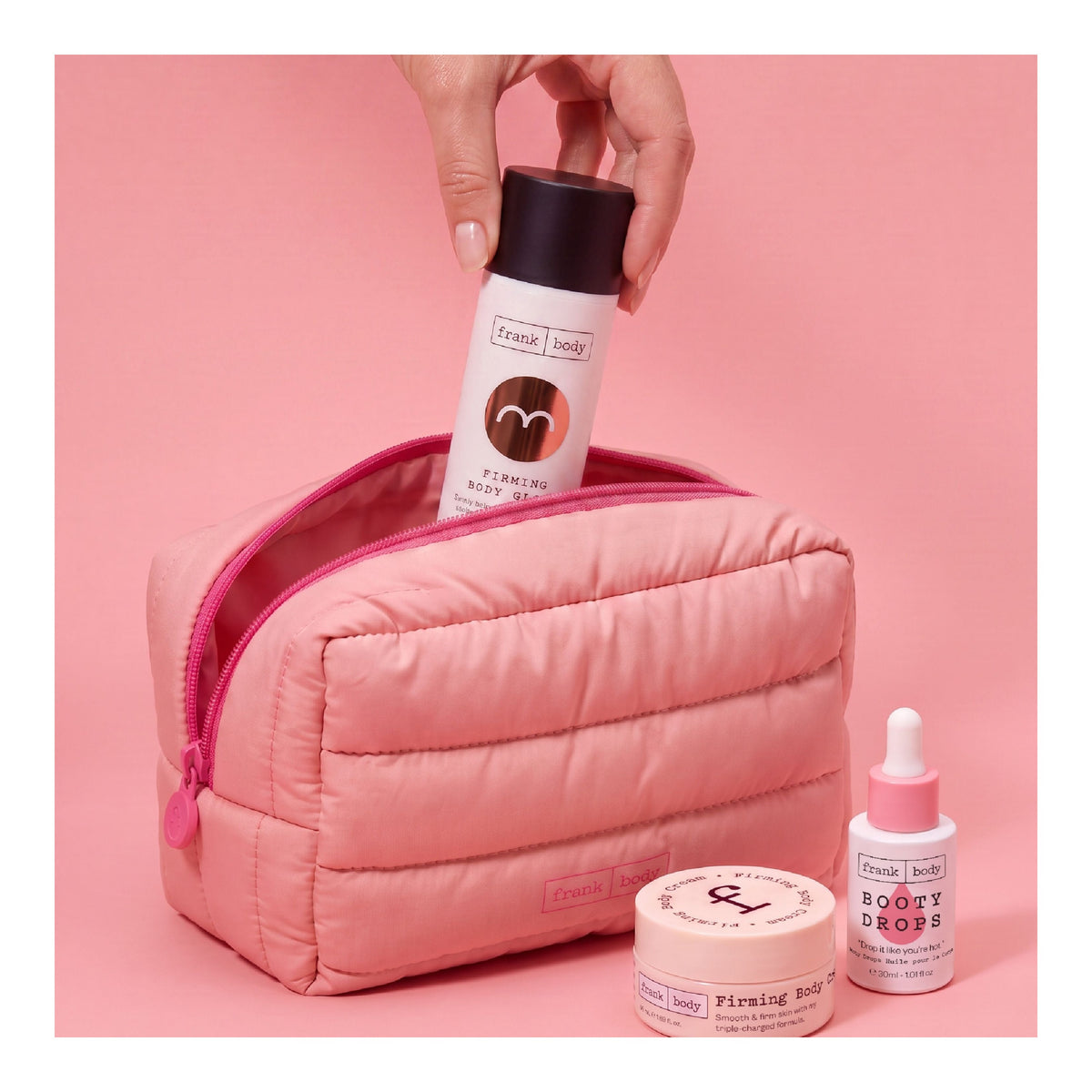Hand steckt FRANK BODY Firming Body Glow in eine rosa Kosmetiktasche neben Booty Drops und Firming Body Cream