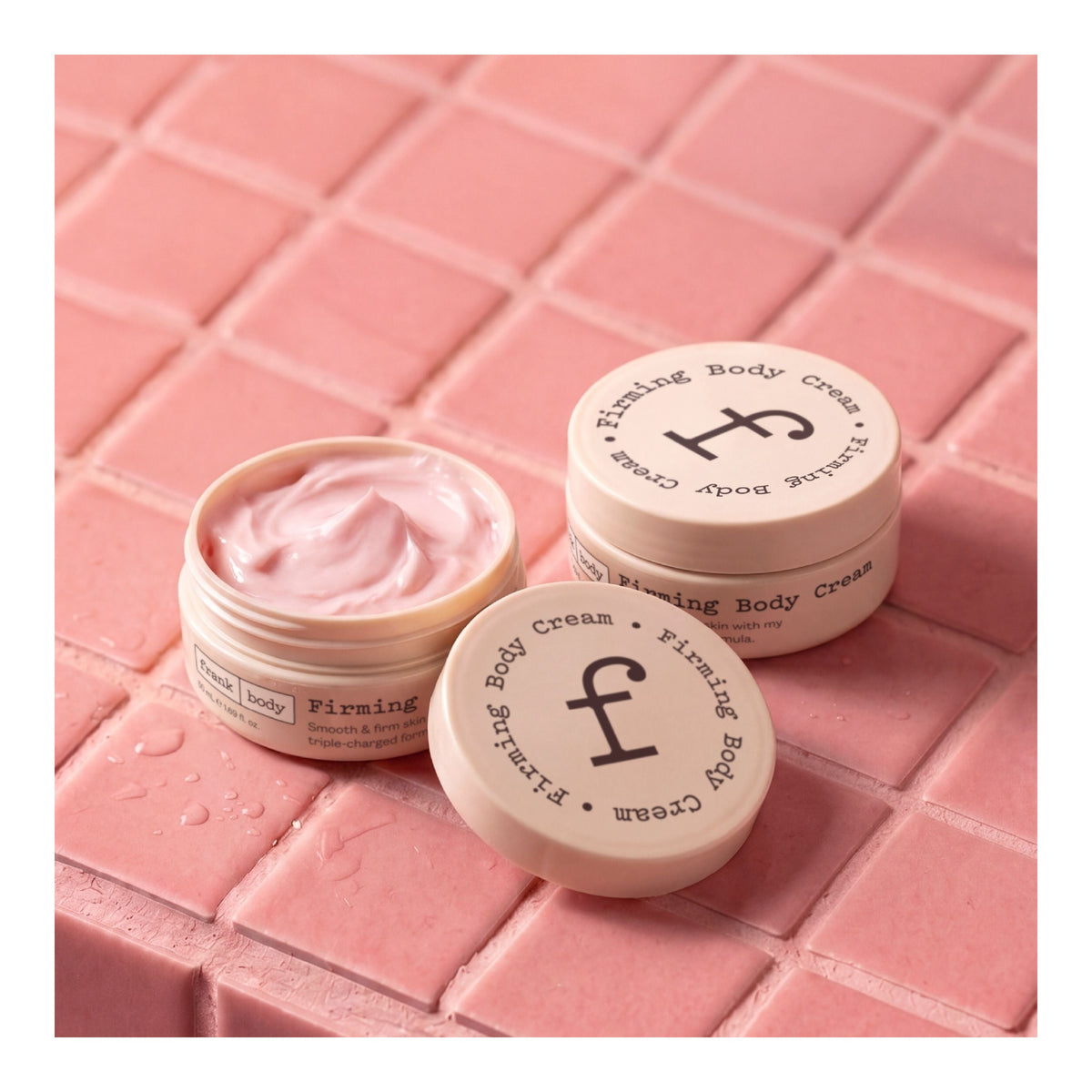 Des pots ouverts et fermés de la crème raffermissante pour le corps FRANK BODY, posés sur un carrelage rose parsemé de gouttes d'eau