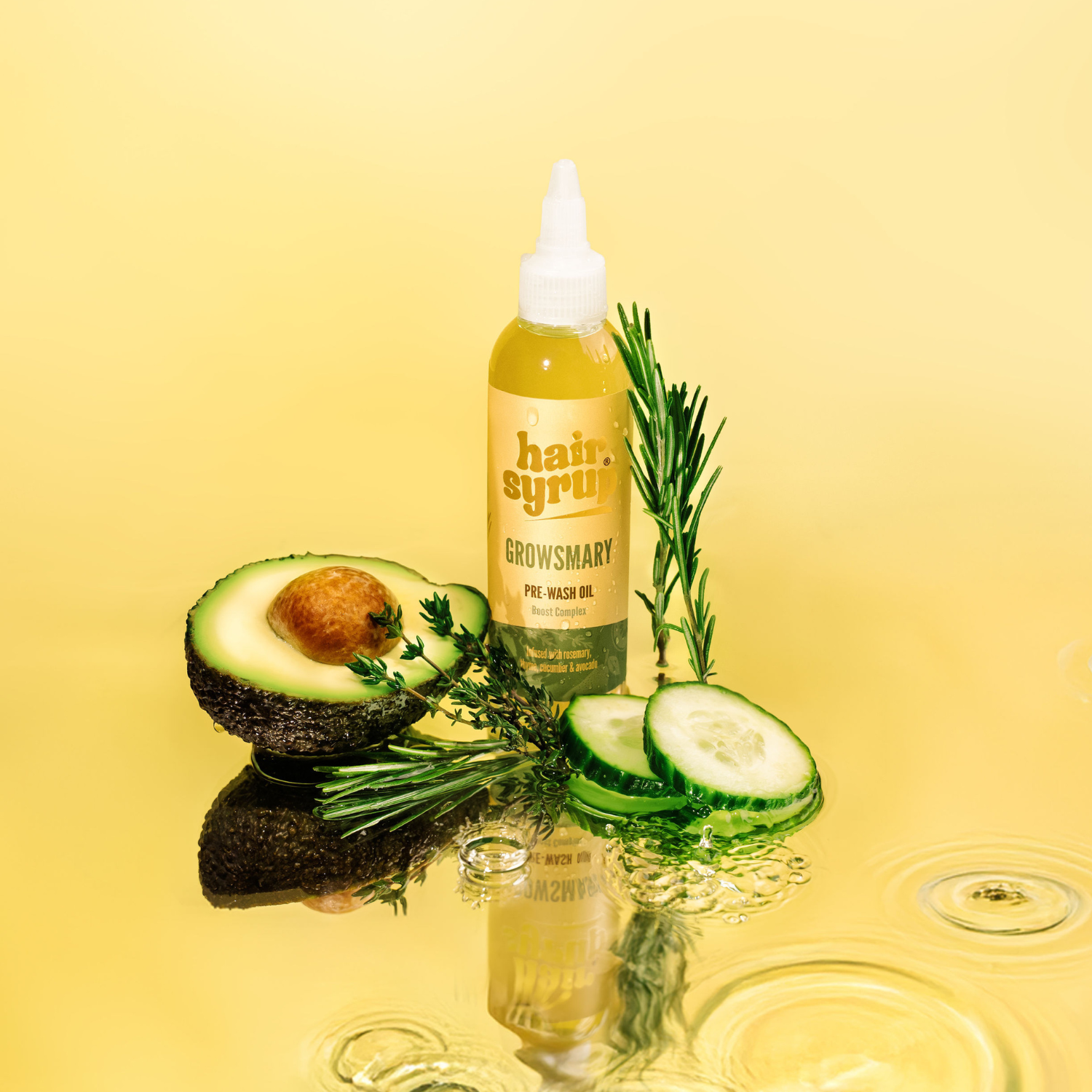 Auf gelbem Grund liegen eine halbe Avocado, Gurkenscheiben, Rosmarin, Thymian und eine Flasche Haaröl mit gelbem Inhalt. Es ist eine spiegelnde Oberfläche mit Wassertropfen zu sehen.