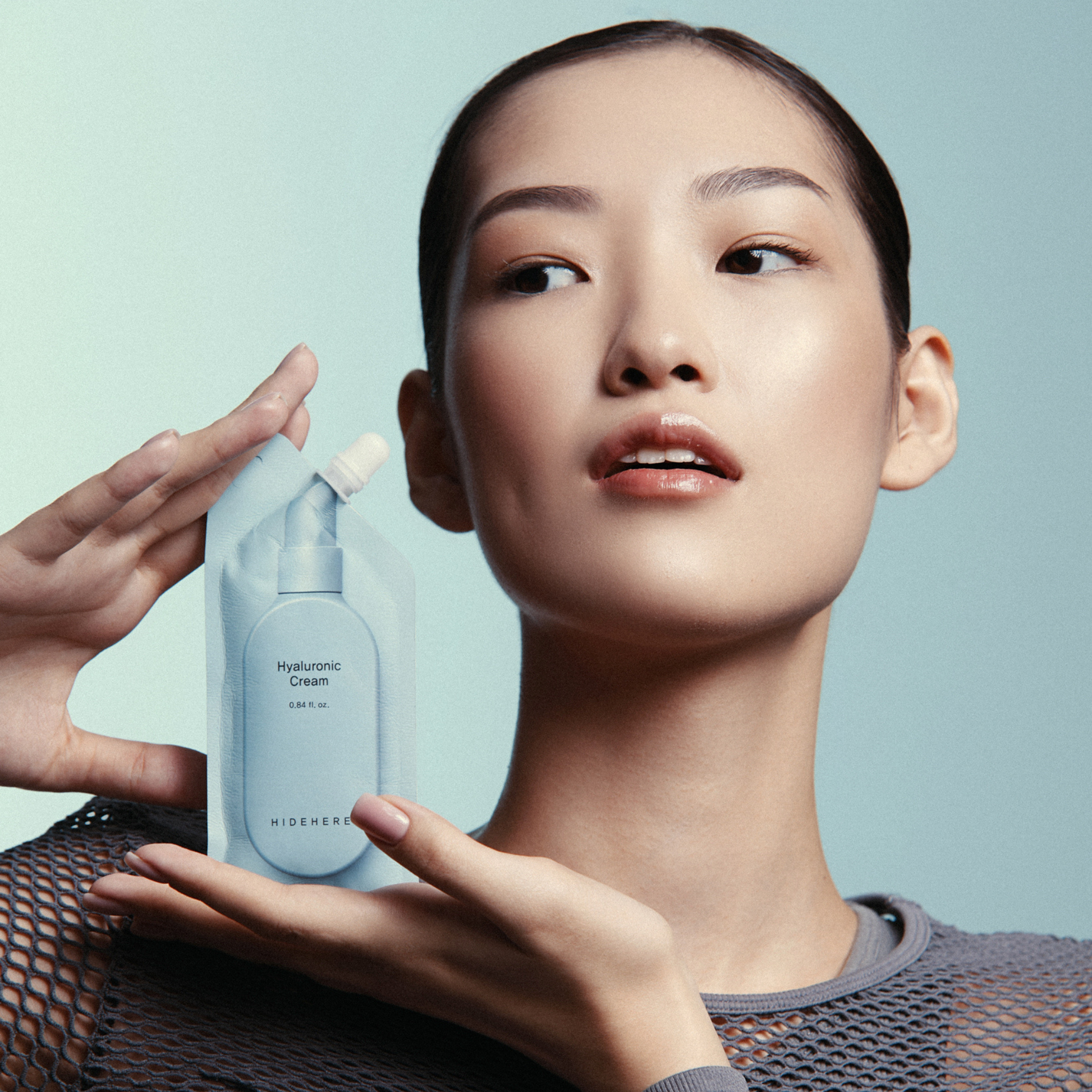 Eine Frau mit geglättetem Haar hält einen hellblauen Beutel mit der Aufschrift "Hyaluronic Cream" und "HIDEHERE" hoch und schaut nach rechts. Sie trägt ein graues Netzoberteil vor einem hellblauen Hintergrund.
