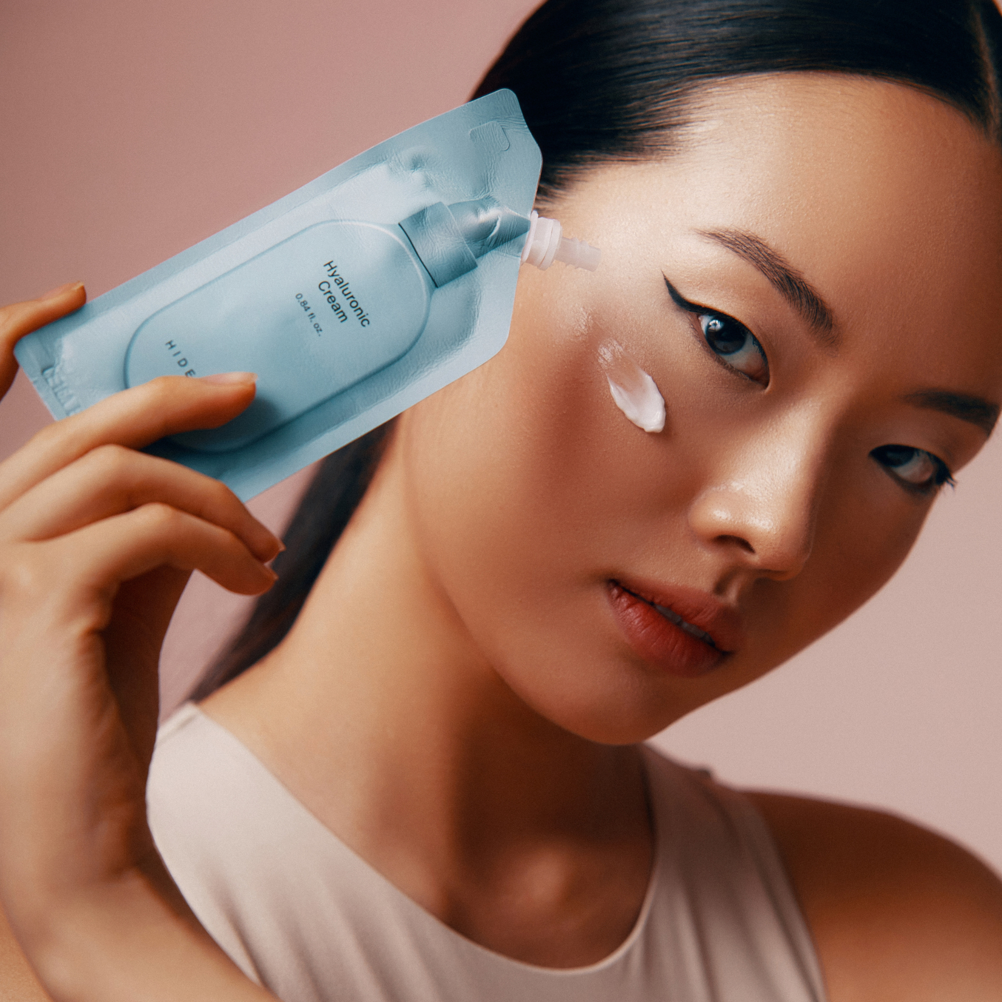 Nahaufnahme einer asiatischen Frau, die eine Hyaluronsäurecreme aus einer hellblauen Packung auf ihr Gesicht aufträgt. Die Packung ist mit "Hyaluronic Cream 0.84 fl.oz." beschriftet. Die Frau blickt mit einem leichten Lächeln nach unten.
