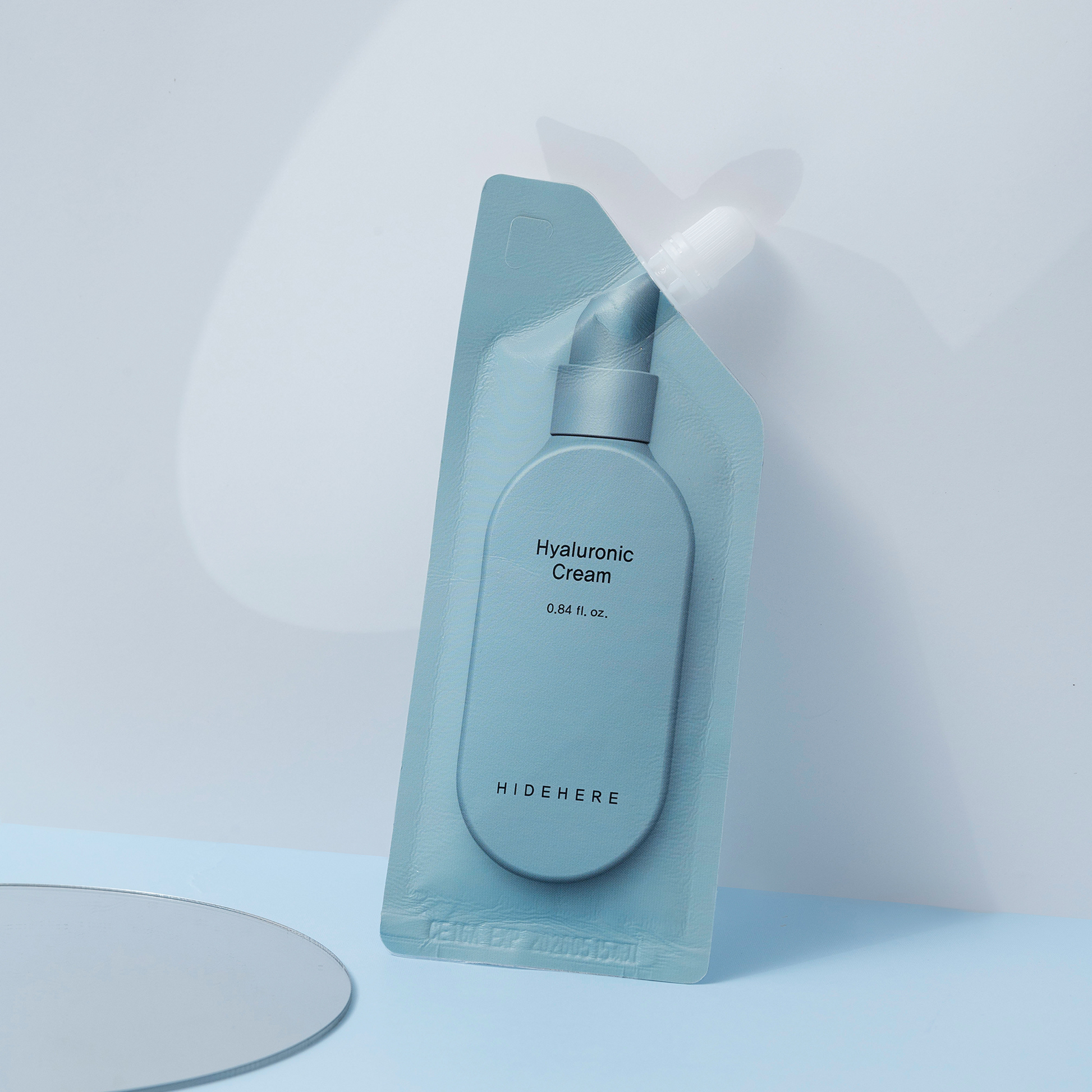 Eine tealfarbene Hyaluronic Cream Probe von HIDEHERE ist in einem tealfarbenen Kunststoffbehälter auf einer hellblauen Oberfläche mit einem Spiegel in der unteren linken Ecke und einem hellblauen Hintergrund verpackt.
