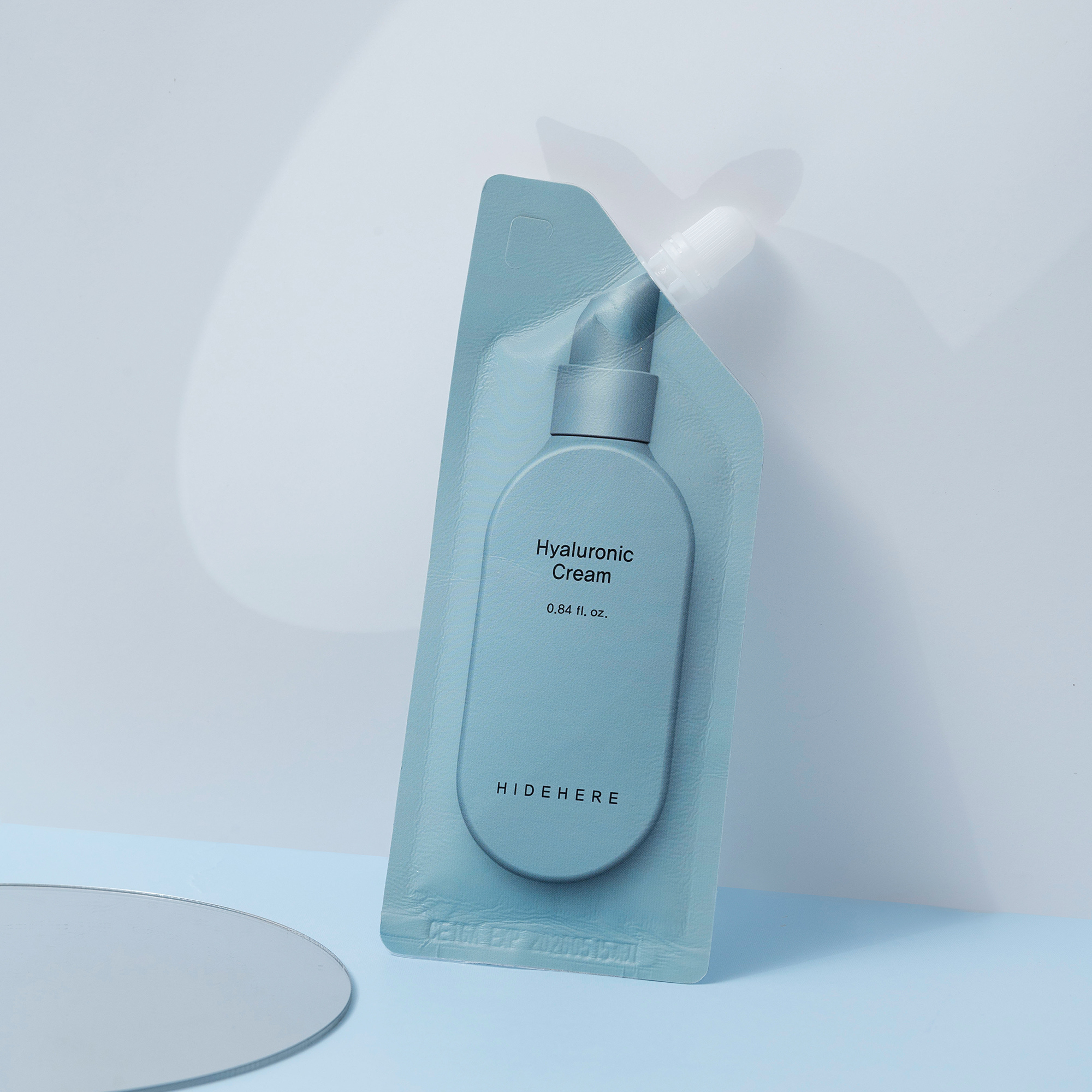 Eine tealfarbene Hyaluronic Cream Probe von HIDEHERE ist in einem tealfarbenen Kunststoffbehälter auf einer hellblauen Oberfläche mit einem Spiegel in der unteren linken Ecke und einem hellblauen Hintergrund verpackt.
