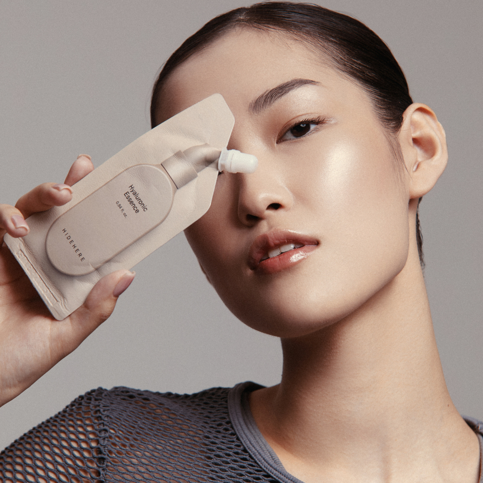 Eine Person mit zurückgegelten dunklen Haaren hält vor einem hellgrauen Hintergrund ein beiges "HIDEHERE Hyaluronic Essence"-Schönheitsprodukt neben ihr Gesicht. Sie trägt ein graues Netzoberteil und blickt ruhig zum Betrachter.
