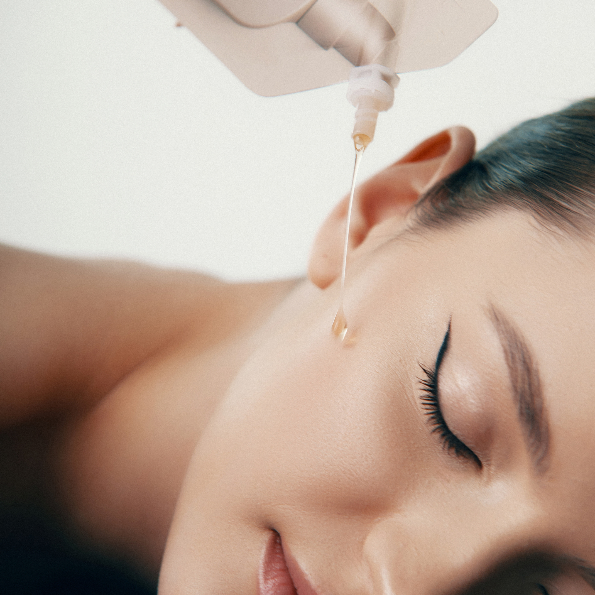 Nahaufnahme des Gesichts einer Frau mit geschlossenen Augen, die mit Öl behandelt wird. Ein Behälter gießt einen Ölstrahl in die Nähe ihres Ohrs. Glatte Haut und elegantes Make-up. Die Szene suggeriert Entspannung und Hautpflege.