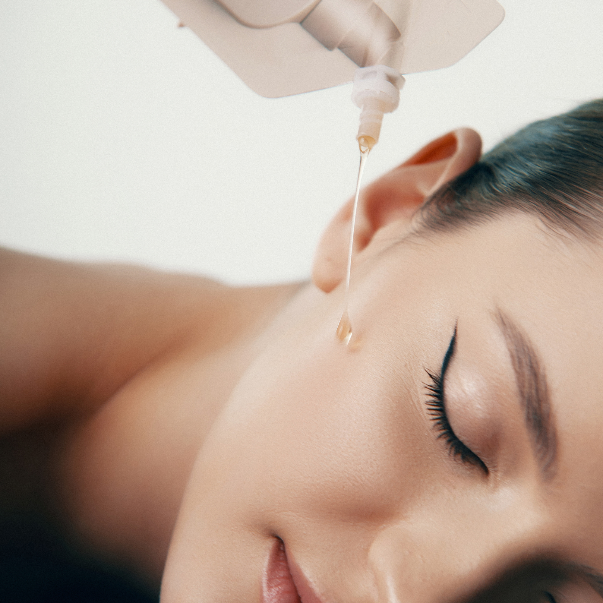 Nahaufnahme des Gesichts einer Frau mit geschlossenen Augen, die mit Öl behandelt wird. Ein Behälter gießt einen Ölstrahl in die Nähe ihres Ohrs. Glatte Haut und elegantes Make-up. Die Szene suggeriert Entspannung und Hautpflege.
