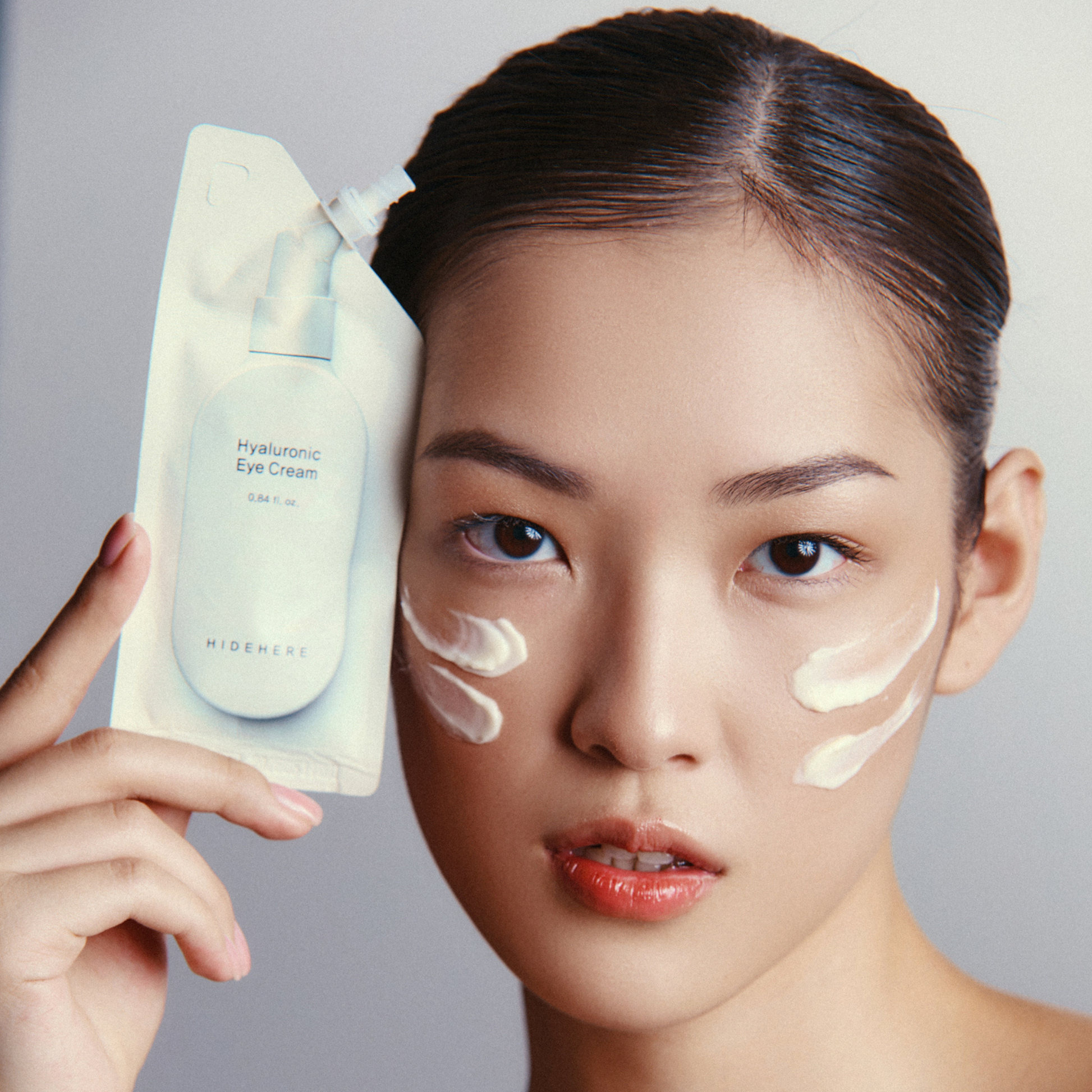 Eine Frau mit hochgestecktem Haar hält eine Packung Hyaluronic Eye Cream von "HIDEHERE" in der Hand. Die Creme wird unter den Augen und auf der Wange aufgetragen.
