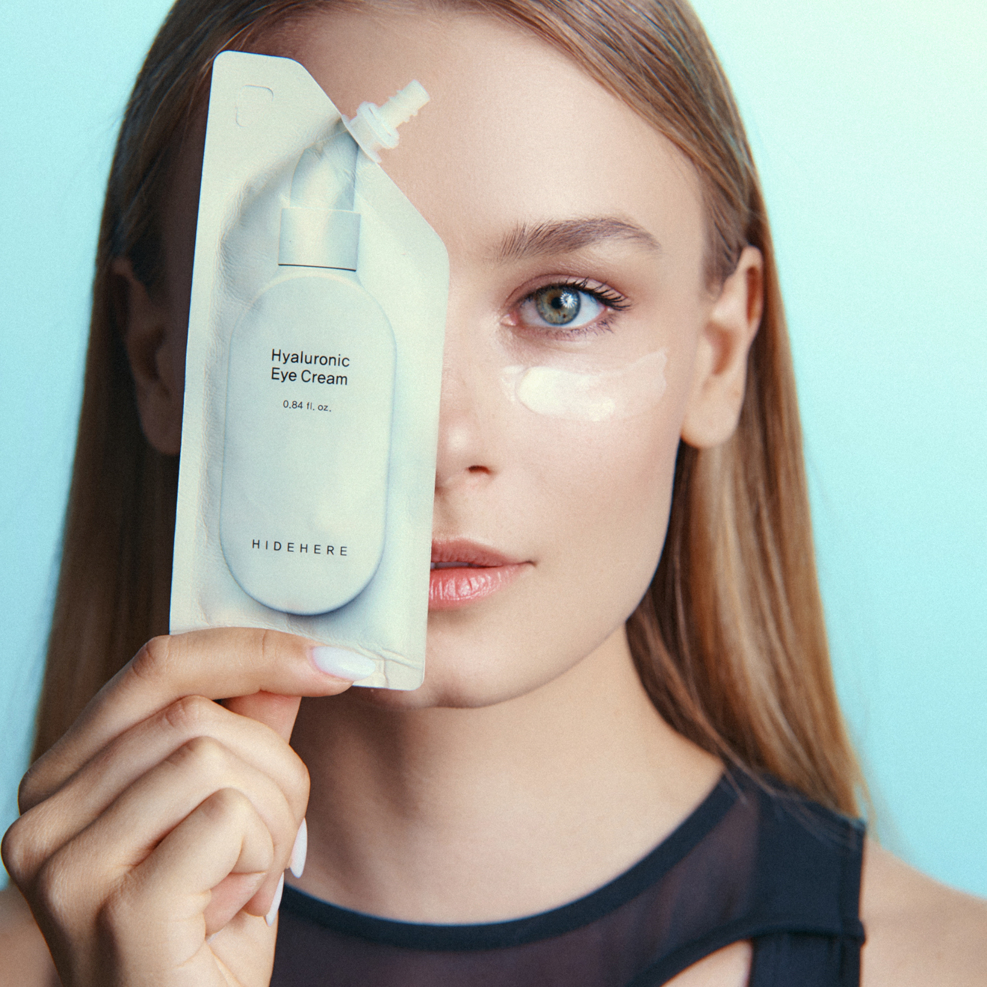 Eine Frau mit einem Klecks Augencreme unter dem Auge hält eine Packung "Hyaluronic Eye Cream" hoch, um die andere Gesichtshälfte abzudecken. Sie hat langes, glattes Haar, helle Haut und trägt ein schwarzes Oberteil. Hellblauer Hintergrund.
