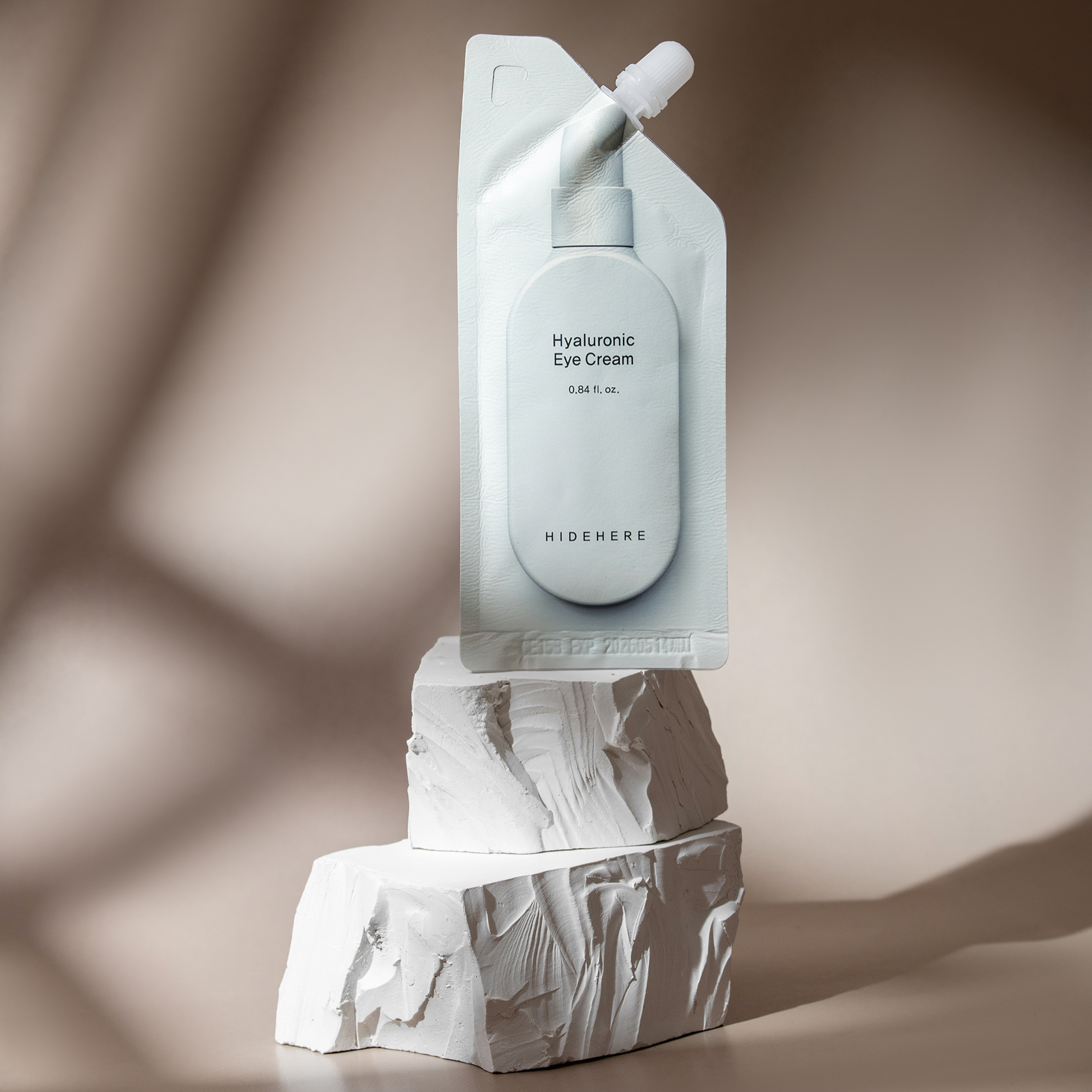 Ästhetische Produktaufnahme: Hyaluronsäure-Augencremebeutel von HideHere, der auf gestapelten weißen Strukturblöcken ruht. Beigefarbener Hintergrund mit weichen Schattenmustern. Minimalistisches Schönheitskonzept.