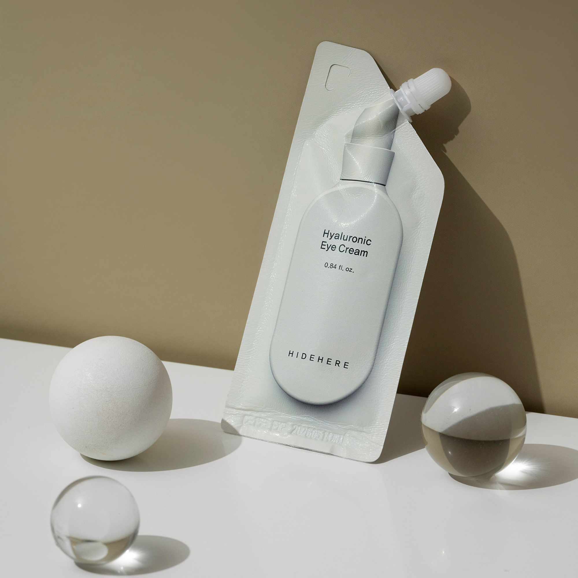 Eine weiße Packung "Hyaluronic Eye Cream" liegt schräg auf einer weißen Fläche, neben einer weißen strukturierten Kugel und zwei klaren Kugeln, vor einem beigen Hintergrund. Die Packung hat einen Schraubverschluss und die Marke "HIDEHERE" aufgedruckt.