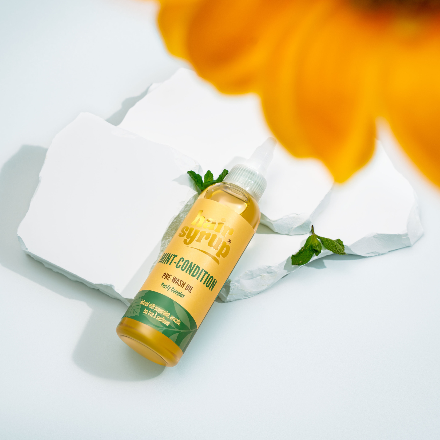 Ein goldfarbenes Ölfläschchen auf weißen Steinen, dekoriert mit Minze, vor einem gelben Blütenblatt auf hellem Grund. Das Etikett trägt goldene Schrift.