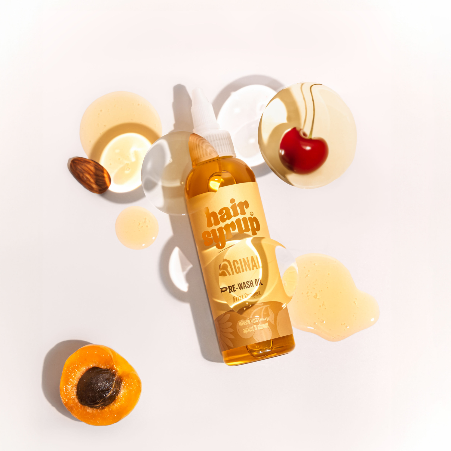 Draufsicht: Haaröl-Flasche mit Mandel, Aprikose, Kirsche und Öltropfen auf weißem Grund.