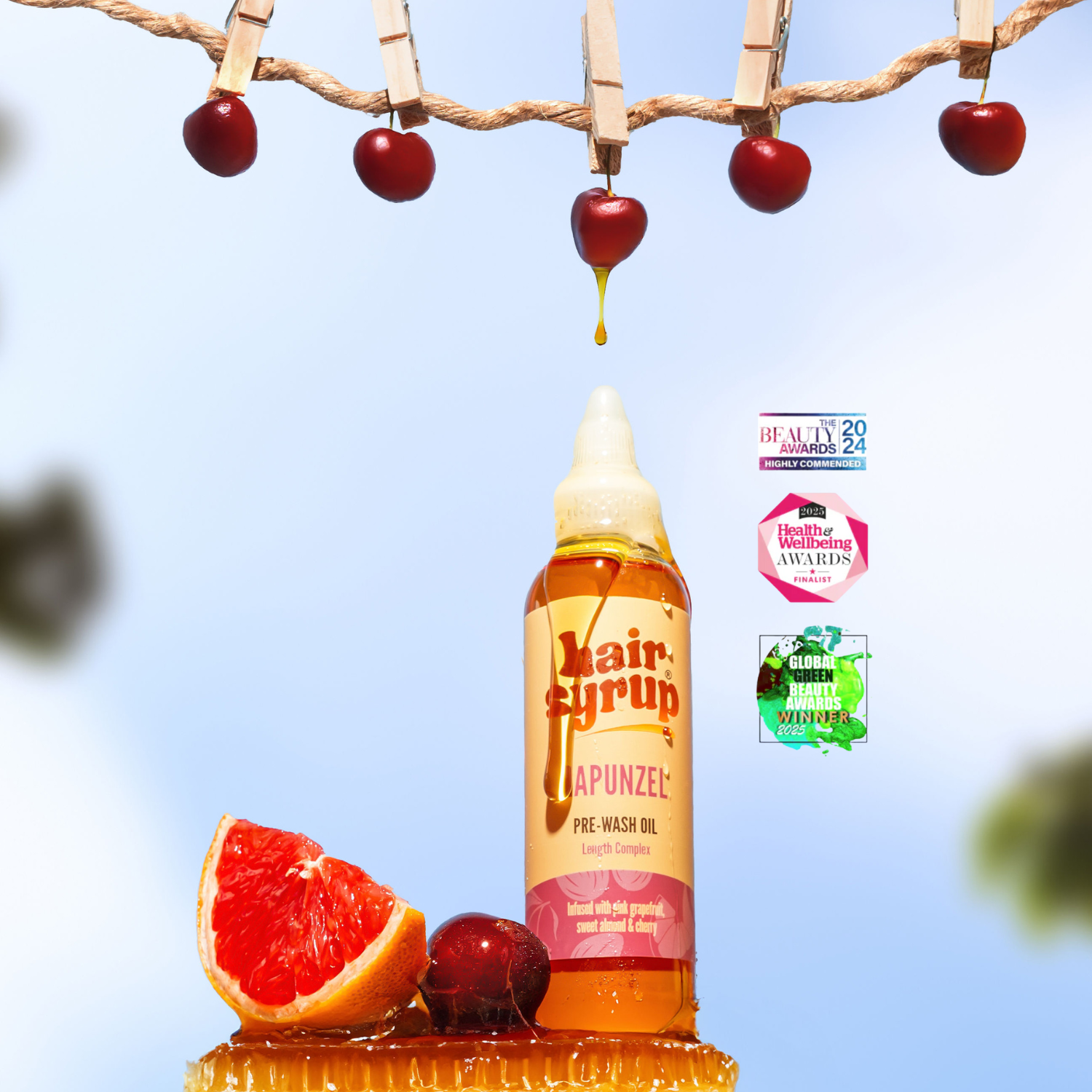 Ein Flasche Haaröl steht auf einer Honigwabe, daneben eine Grapefruit und Kirsche. Darüber hängen Kirschen an einer Leine. Aus einer Kirsche tropft Öl. Mehrere Auszeichnungen sind im Hintergrund.