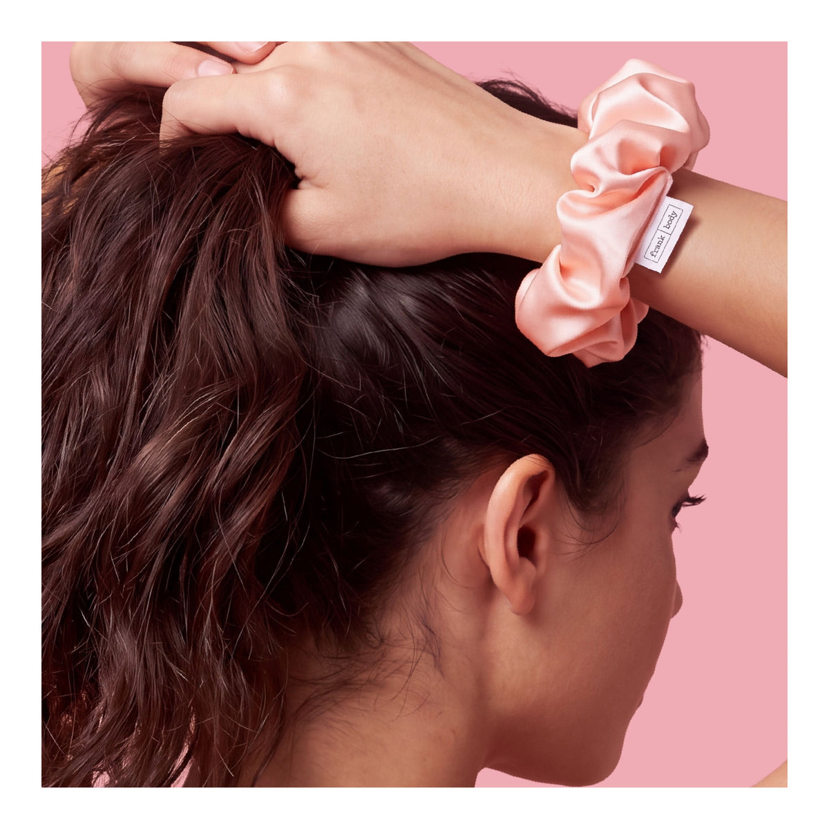 Eine Frau greift in ihr braunes Haar, am Handgelenk ein rosa Satin-Haargummi von FRANK BODY vor rosa Hintergrund