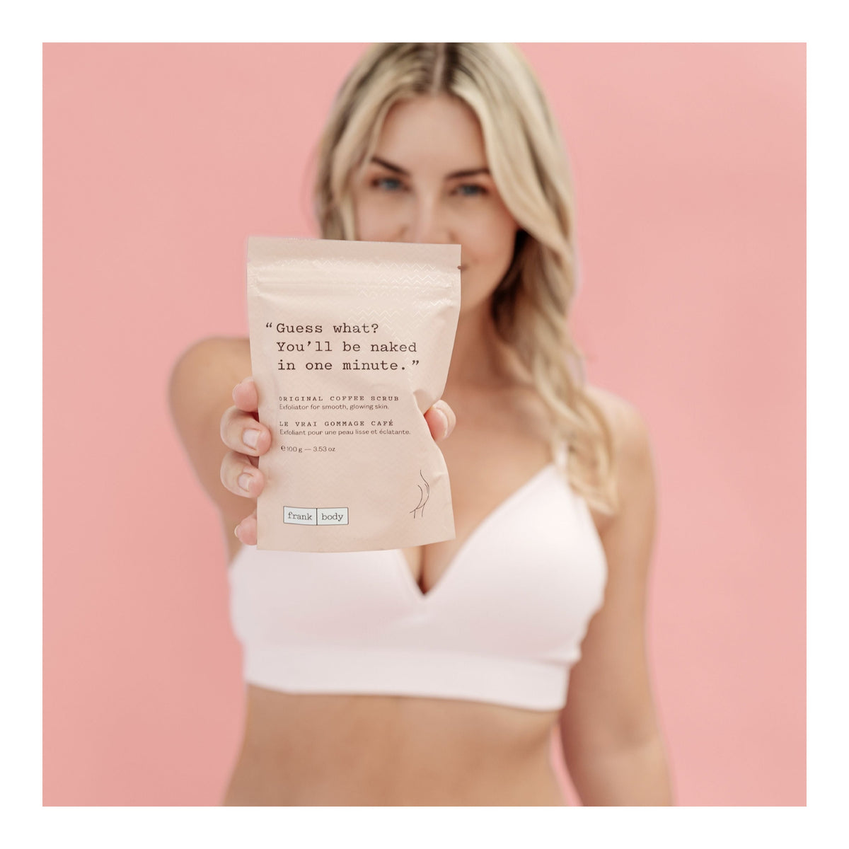 A woman holds the coffee scrub from the FRANK BODY Triple Shot Kit up to the camera against a pink background