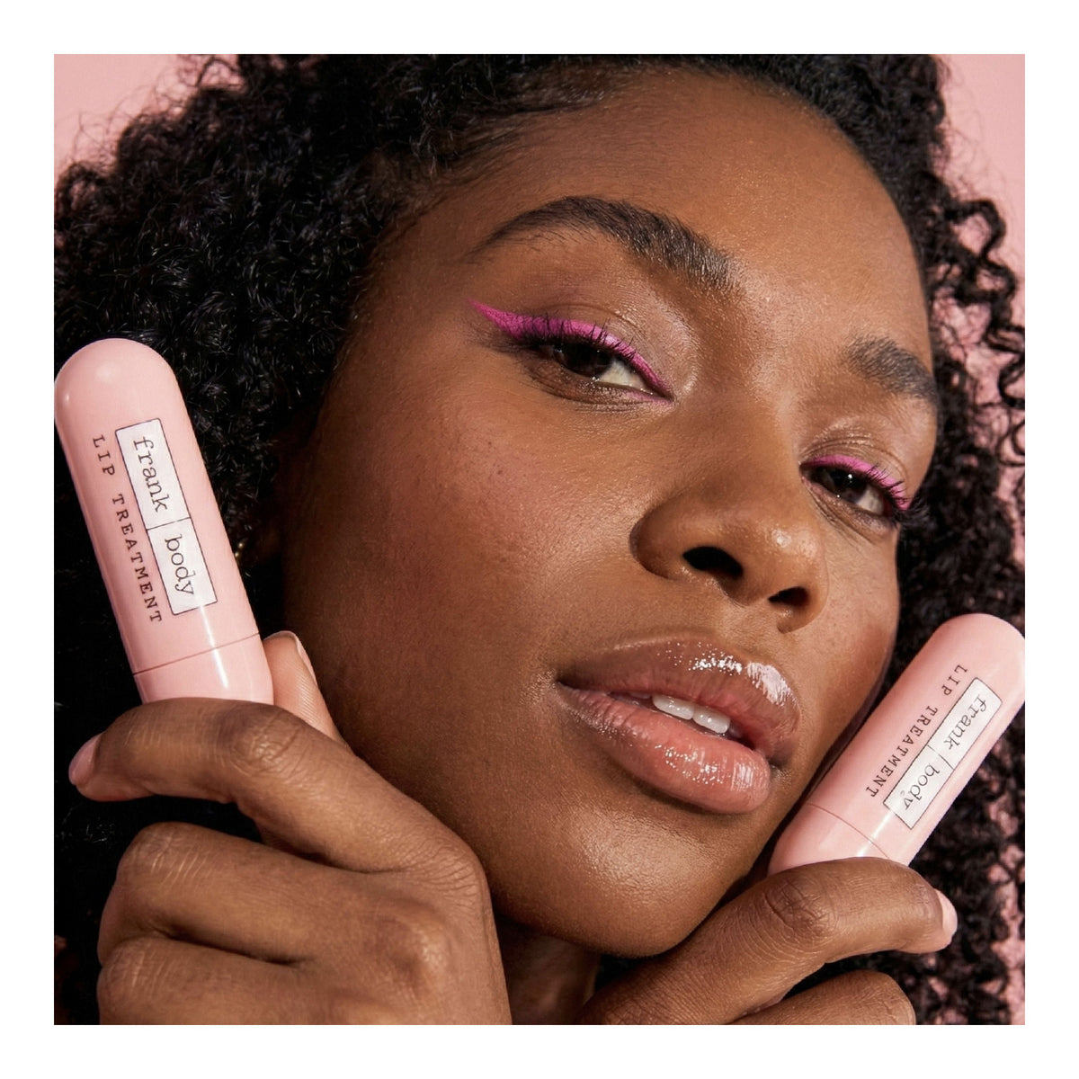 A woman with pink eyeliner holds two pink FRANK BODY Lip Treatment sticks next to her glossy lips