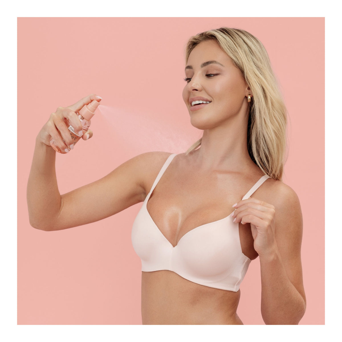 A blonde woman wearing a light-colored bra sprays FRANK BODY spray on her cleavage against a pink background