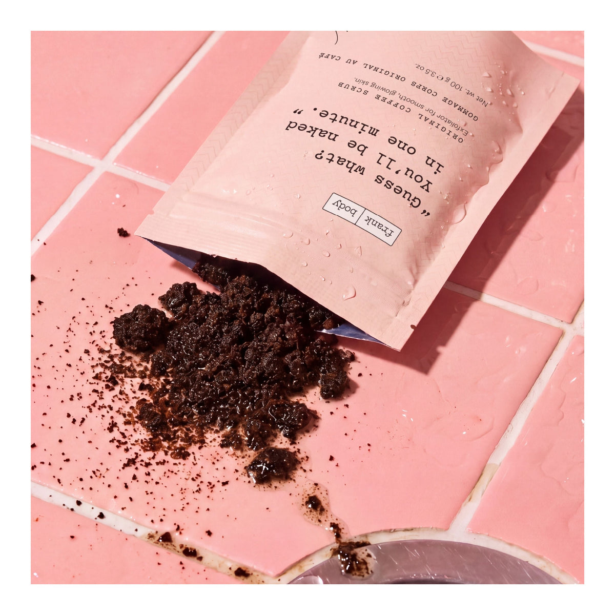 Dunkles FRANK BODY Kaffeepeeling, verschüttet auf feuchten, rosa Badezimmerfliesen neben der Produktverpackung