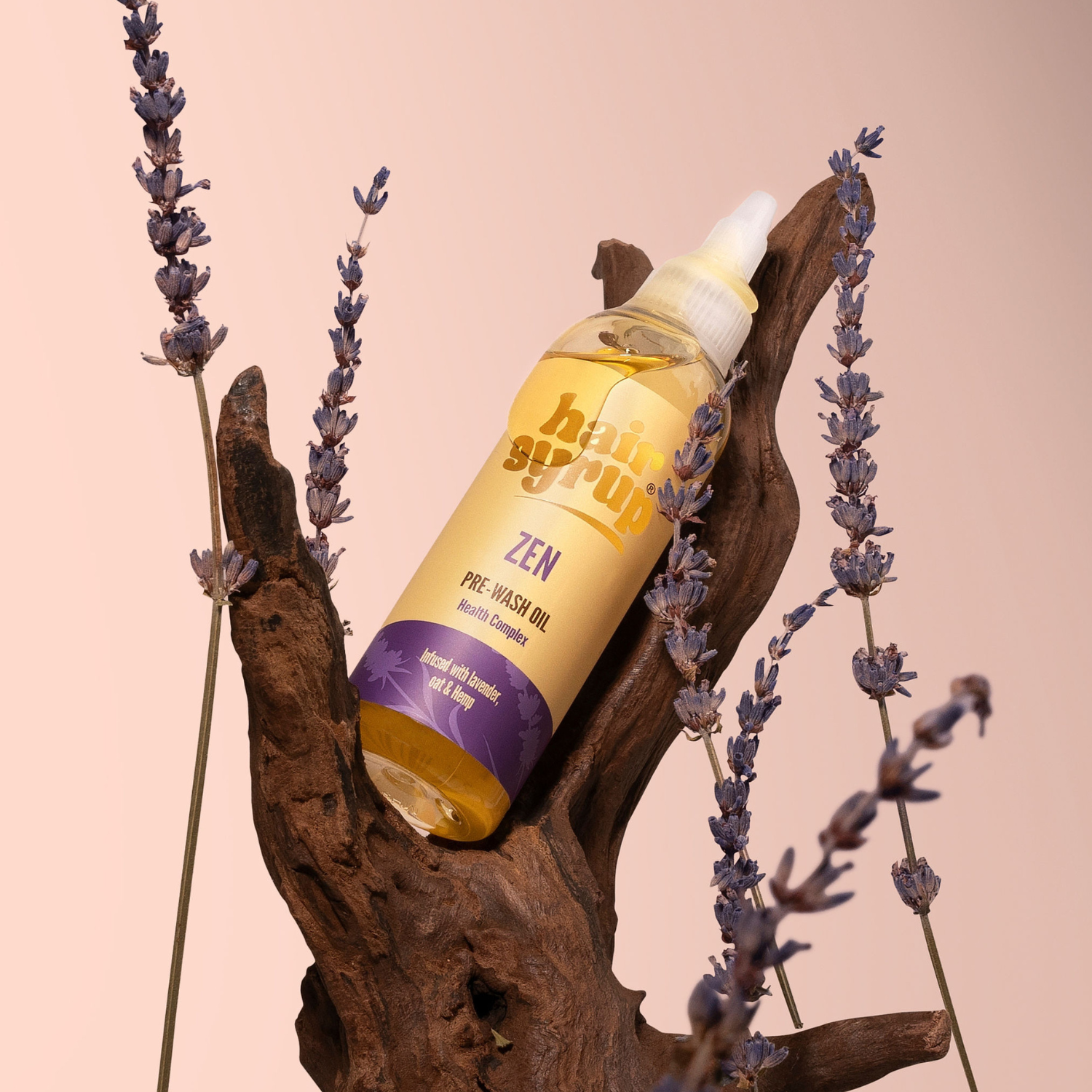 Eine Flasche Haaröl mit Lavendel steht auf einem Stück Holz, umgeben von Lavendelzweigen. Das Öl ist gelb und die Etikette ist gold und violett.