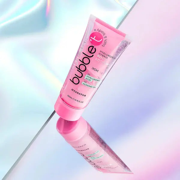 Eine Tube rosa Bubble T Hybrid Body Lotion Hyaluronic liegt auf spiegelndem Untergrund. Sie enthält Hyaluronsäure und Mandelöl. Der Hintergrund ist schillernd.