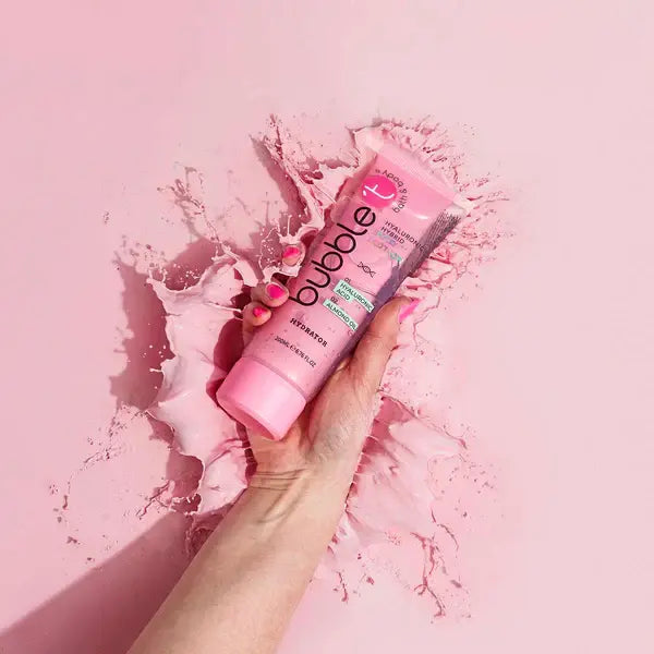 Eine Hand hält eine rosa "Bubble T Hybrid Body Lotion Hyaluronic". Der Hintergrund ist rosa mit Farbspritzern.