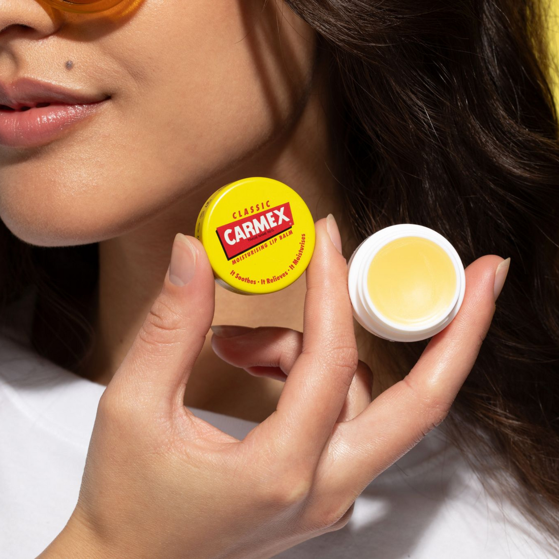 Frau hält zwei CARMEX Classic Lippenbalsam Tiegel. Einer ist geschlossen, einer offen. Gelber Hintergrund.