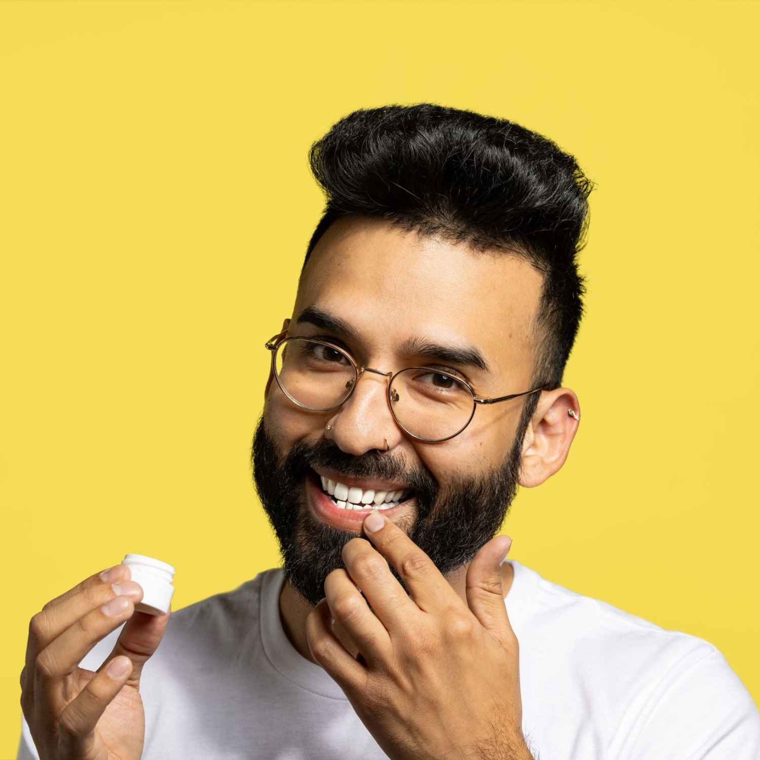 Mann mit Brille, der Lippenbalsam aus einem Tiegel auf seine Lippen aufträgt. Gelber Hintergrund.