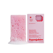 Rosa Schwammseife mit Verpackung. Die Seife ist rechteckig mit einer unebenen Oberseite und hat eine schaumige Basis. Die Verpackung ist rosa mit Text.