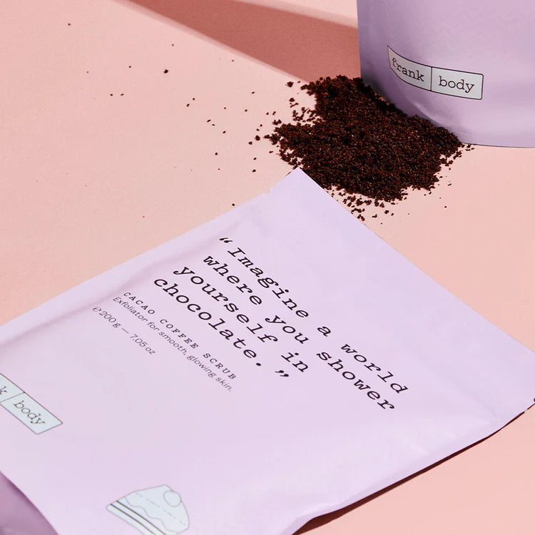 Fliederfarbene Verpackung des FRANK BODY Cacao Coffee Scrub neben dunklem Kaffee-Peeling auf rosa Hintergrund