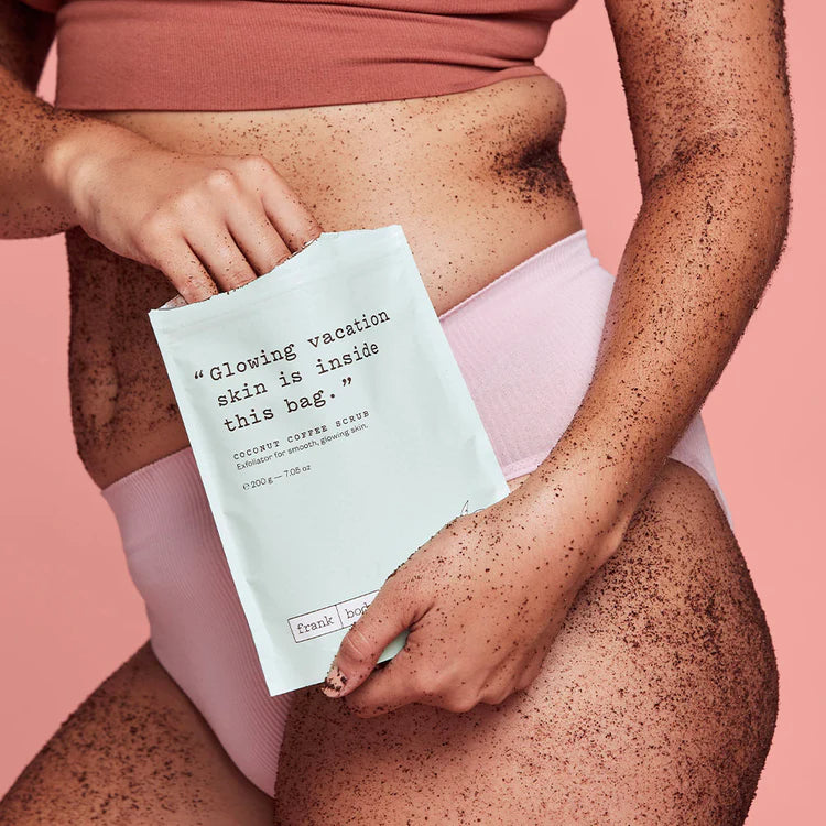 Person mit Kaffeepeeling auf der Haut hält eine Packung FRANK BODY Coconut Coffee Scrub vor rosa Hintergrund