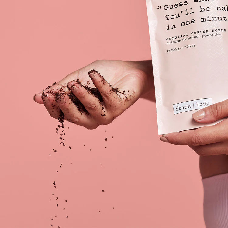 Kaffeesatz rieselt aus einer Hand neben der Verpackung des FRANK BODY Original Coffee Scrub vor rosa Hintergrund