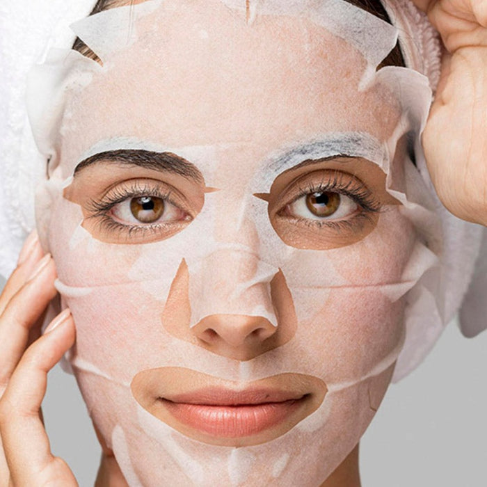 Nahaufnahme einer Frau mit Tuchmaske, die Partien für Augen, Nase und Mund freilässt. Die Maske soll die Haut mit Jasmin, Aloe Vera & Niacinamid pflegen.